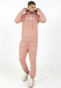 Felpa rosa con tasca frontale, abbinata a pantaloni jogger coordinati. Entrambi presentano il nome del marchio "SIXTHJUNE" in bianco. Completano l'outfit delle sneakers marroni.