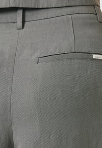 Pantaloni grigi con una texture liscia, vestibilità slim e una tasca posteriore con un'etichetta piccola. Design sottile con dettagli di cucitura minimalisti.