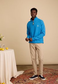 Blauwe fleece pullover met halve ritssluiting en logo, gecombineerd met beige broek en donkere sneakers op een patroonrijke tapijt tegen een neutrale achtergrond.