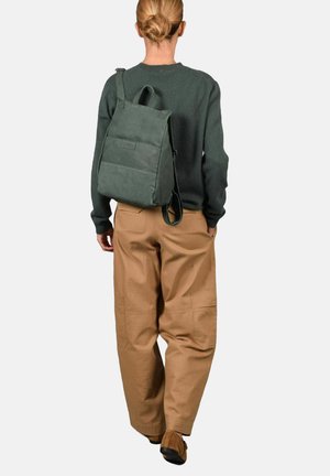 Mochila verde con textura, cierre en la parte superior enrollado y correa ajustable, llevada por una persona con un suéter gris y pantalones beige.