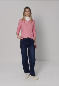 Top color rosa chiaro a maniche lunghe con scollo con zip, abbinato a jeans a gamba larga blu scuro e sneakers bianche, su sfondo semplice.
