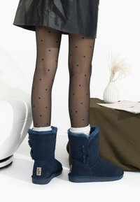 Botines de ante azul marino con un suave forro de piel de pelo azul, que presentan dos hebillas decorativas, combinados con medias negras con lunares y una falda negra.