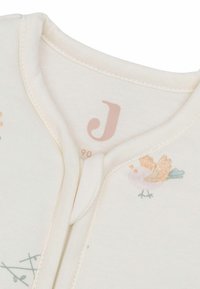 Un grenouillère en coton de couleur crème avec une broderie d'oiseau pastel, fermeture pression et encolure arrondie. Présente une texture douce et une lettre imprimée subtile.