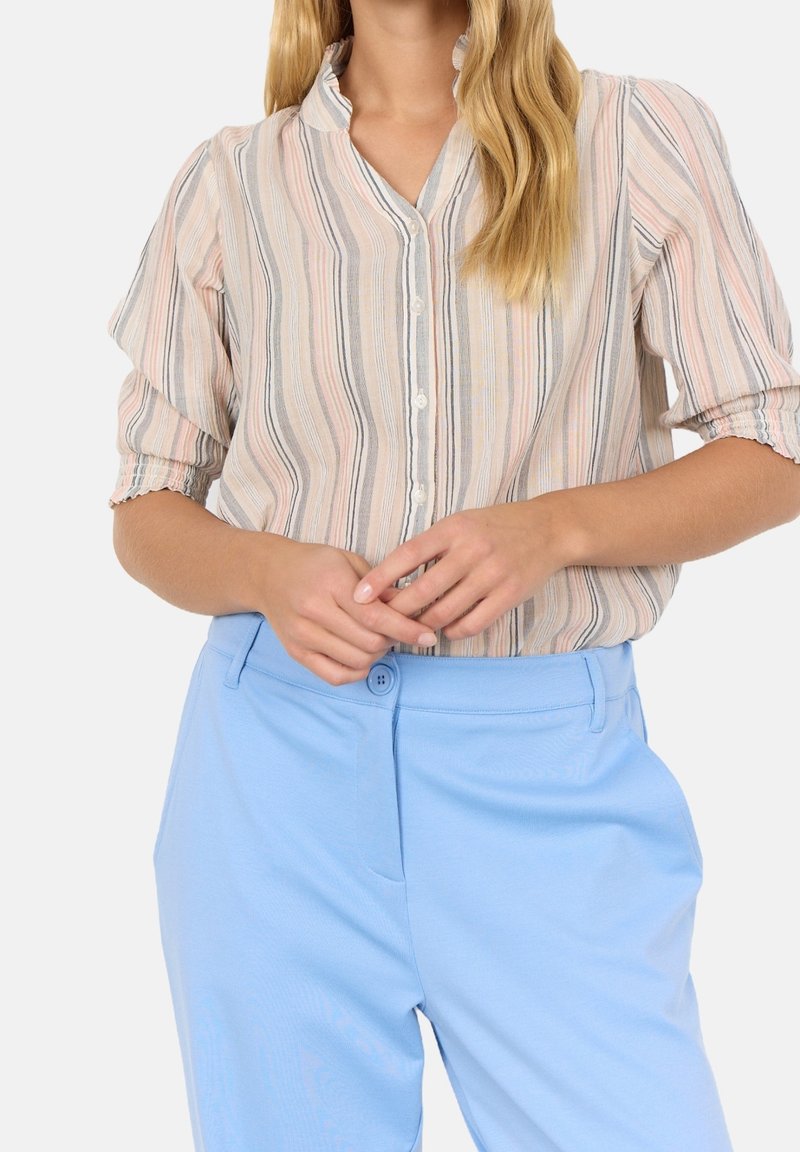 Femme portant une blouse à rayures pastel avec manches bouffantes et un pantalon taille haute bleu clair, mains jointes près de la taille.