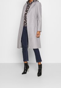Manteau gris en mélange de laine avec un design à double boutonnage, associé à un sweat-shirt noir à imprimé floral, un jean bleu foncé raccourci et des bottines noires à talons.
