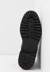 Semelle de botte en caoutchouc noir avec des crampons profonds, positionnée verticalement contre un coin de mur blanc, affichant la taille 37 embossée.