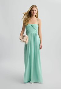 Maxi dress verde menta con spalline sottili, dettaglio a nodo attorcigliato sul busto e gonna fluida. Il modello tiene una pochette con dettagli in piuma.