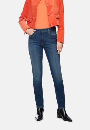 Frau trägt dunkelblaue Jeans mit kleinen Nieten an den Taschen, schwarze Stiefel mit Absatz, orangefarbenes Oberteil und orangene Cordjacke mit Knöpfen.