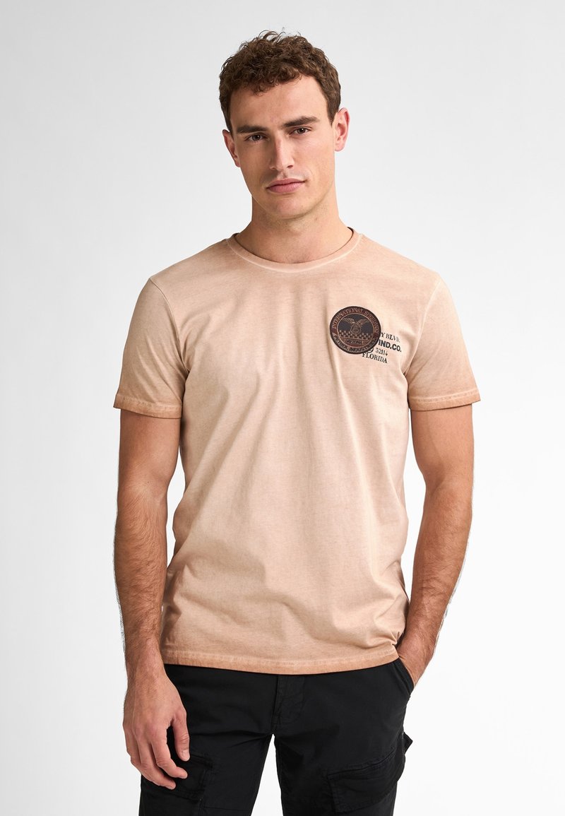 Svetle béžové tričko s krátkým rukávem, na levé straně hrudi s kruhovým logem, s kulatým výstřihem a měkkou texturou látky.