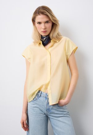 Mujer rubia con una blusa de manga corta de color amarillo claro, jeans azul claro y un pañuelo negro con estampado en el cuello, de pie con una mano en el bolsillo.