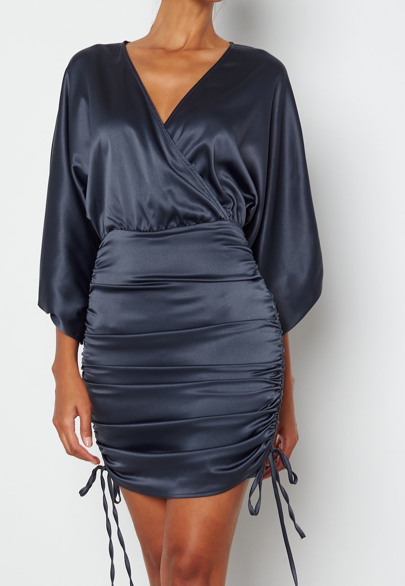 Robe portefeuille en satin bleu marine avec côtés froncés ajustables, col en V et manches longues amples sur une personne debout.