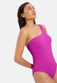 Mujer de cabello largo y oscuro que lleva un traje de baño magenta texturizado, de un solo hombro, mirando hacia arriba con las manos detrás de la espalda.
