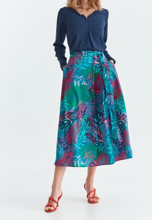 Femme portant un chemisier bleu marine à manches longues, une jupe midi à imprimé tropical coloré avec des poches latérales, et des sandales rouges à talons et brides.