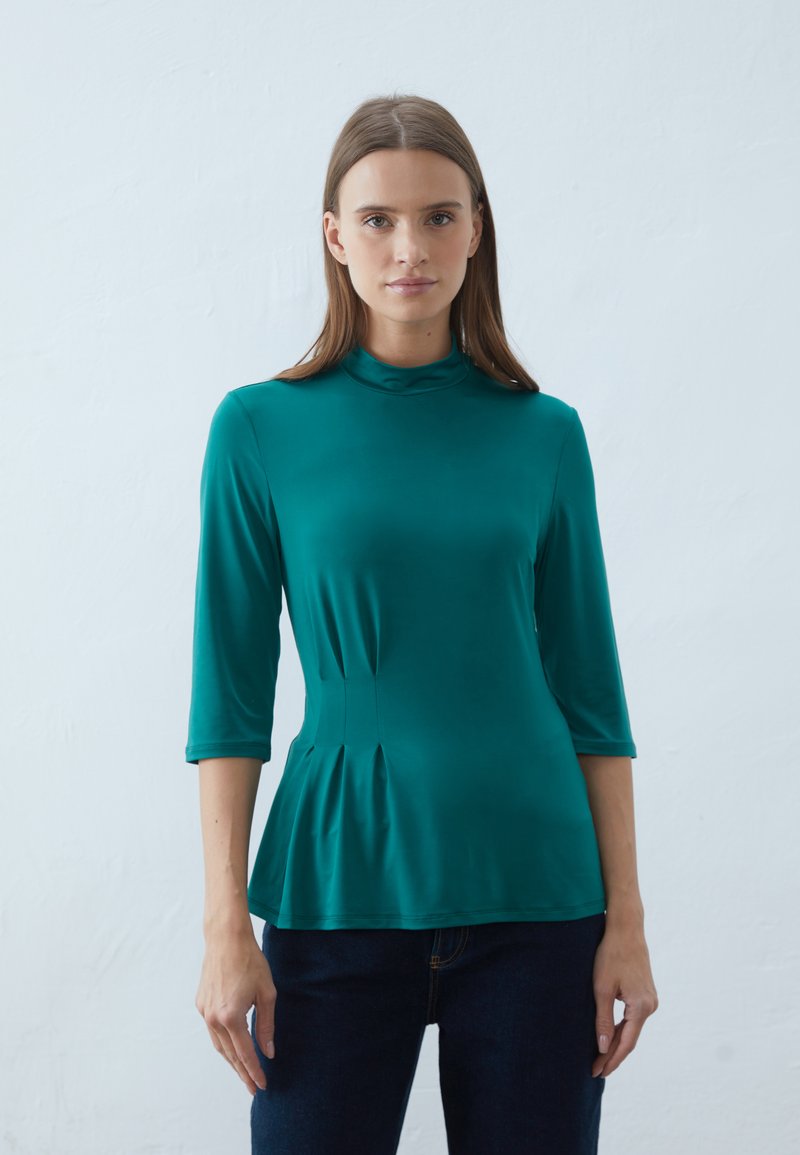 Teal peplum topp med høy hals og trekvartlange ermer, laget av glatt, elastisk stoff. Har subtile plisseringer i midjen.