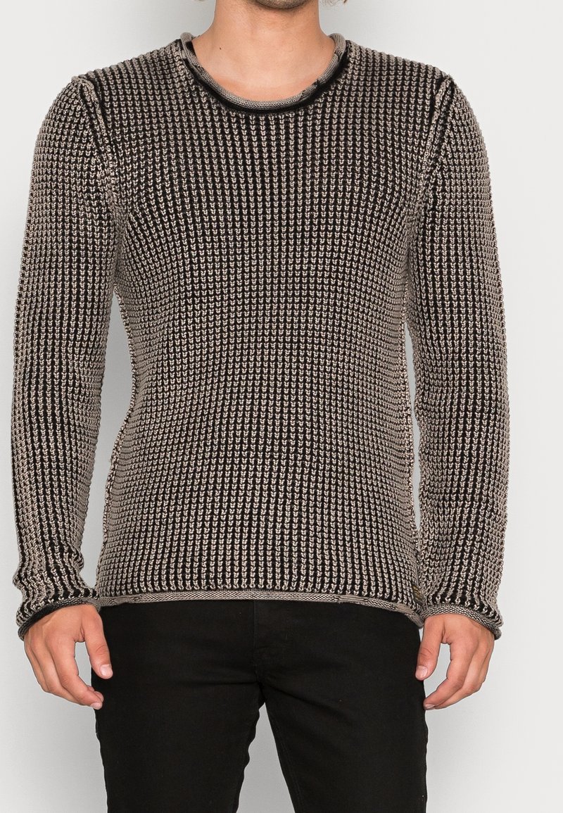 Homme portant un pull en maille texturée noir et beige à manches longues et un pantalon noir sur un fond uni.