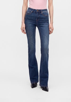 Kvinde iført mørkeblå højttaljede bootcut-jeans med forlommer, kombineret med sorte spidse sko og en lyserød top.