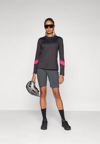 Langärmliges schwarzes Sportoberteil mit pinken Akzenten, kombiniert mit grauen Shorts. Die Accessoires umfassen schwarze Socken, eine Sonnenbrille und einen grauen Helm.