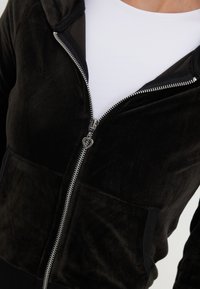 Zwarte fluwelen hoodie met ritssluiting, een zachte textuur, een kangoeroezak en zilveren ritsdetails. Gedragen over een wit shirt.