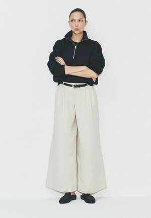 Nainen, jolla on tummat hiukset, musta vetoketjullinen villapaita, kermaan vivahtavat leveälahkeiset housut, musta vyö ja mustat slip-on-kengät, seisoo käsivarret ristissä valkoista seinää vasten.