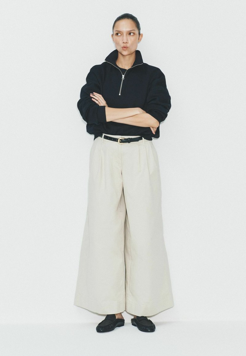 Donna con capelli scuri in maglione nero con zip, pantaloni crema a gamba larga, cintura nera e scarpe nere senza lacci, in piedi con le braccia incrociate contro un muro bianco.