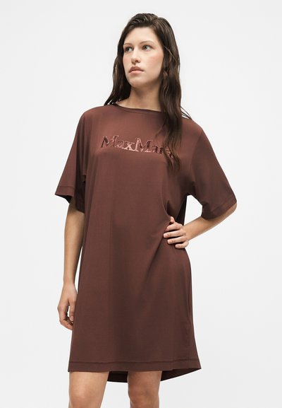 Femme aux longs cheveux foncés portant une robe marron ample avec le logo gaufré "Max Mara", debout avec une main sur la hanche sur fond blanc.