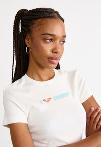 T-shirt à manches courtes couleur crème avec un graphique "I ❤️ SURF" en orange et bleu clair. Fabriqué à partir de matériaux doux avec une coupe ajustée.