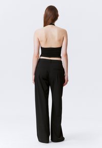 Woman with long brown hair wearing a black halter top and black wide-leg trousers, standing against a plain white background facing away.