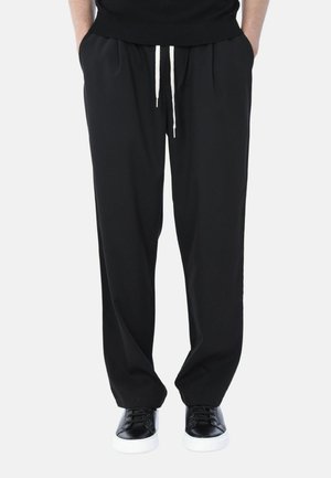 Persona che indossa pantaloni neri larghi con laccetti bianchi, sneaker nere con suola bianca, in piedi con le mani nelle tasche.