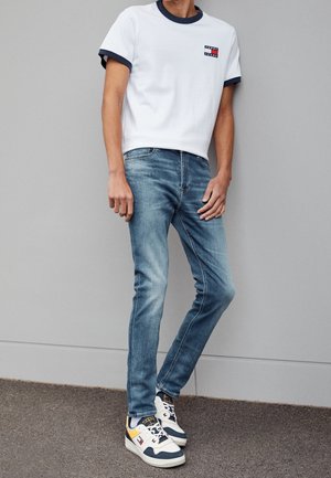 Maglietta bianca con collo blu navy e piccolo logo, jeans slim fit azzurri, sneaker multicolori con accenti gialli e blu navy.