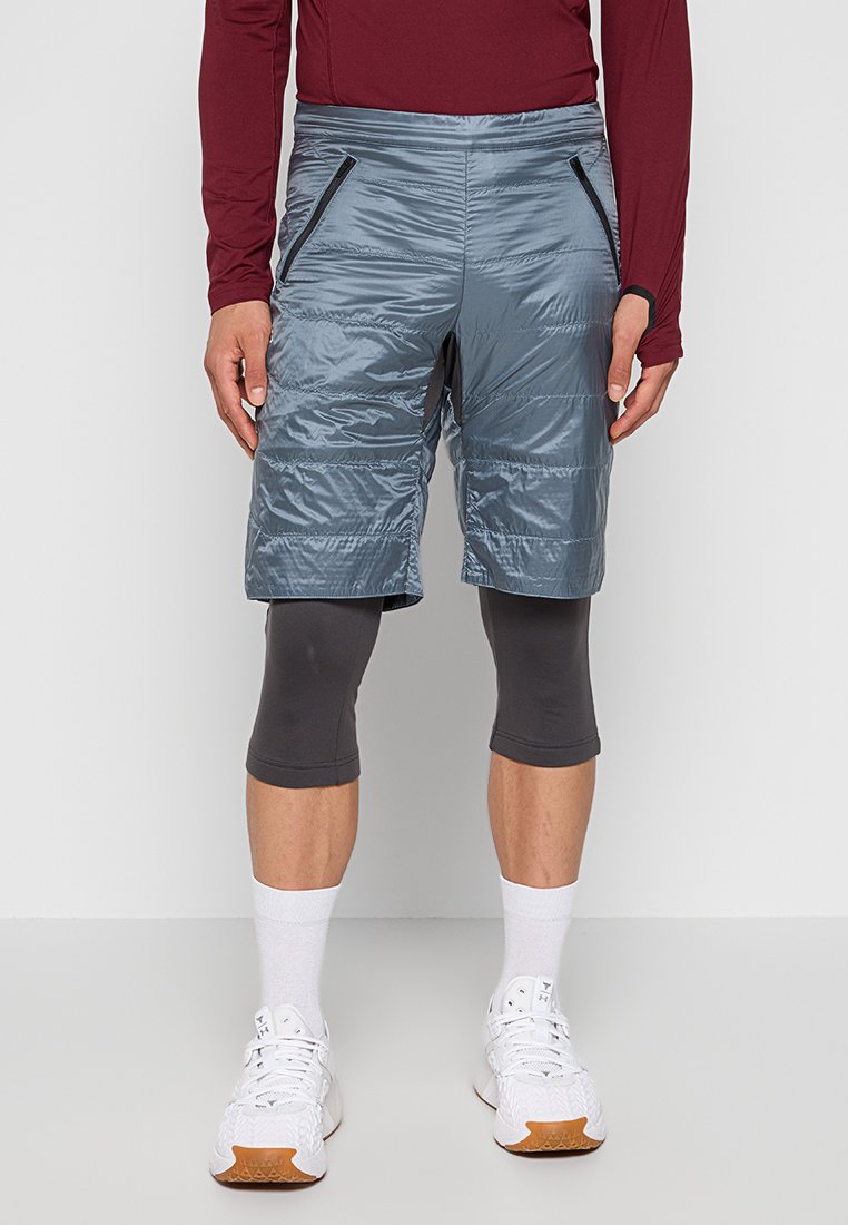 Grå isolerade shorts med en glänsande yta, utrustade med två dragkedjefickor på sidorna och svarta accenter, ihop med tighta grå leggings och vita sneakers.