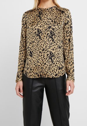 Blouse beige à manches longues avec un motif abstrait noir tacheté et étoilé, portée avec un pantalon en cuir noir, présentée du cou aux hanches.