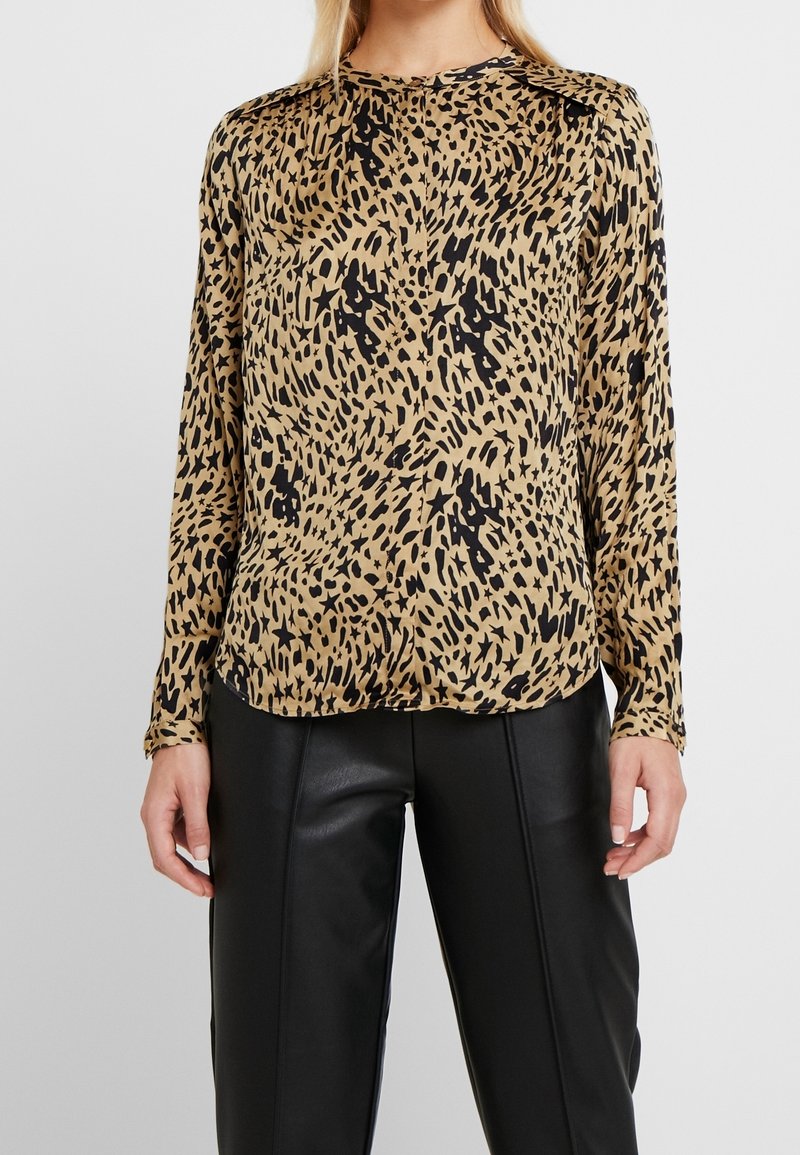 Blouse beige à manches longues avec un motif abstrait noir tacheté et étoilé, portée avec un pantalon en cuir noir, présentée du cou aux hanches.