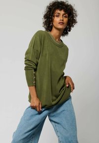 Maglione verde oliva con scollatura rotonda, polsini a coste con bottoni dorati, abbinato a jeans a gamba larga azzurri. Texture liscia.
