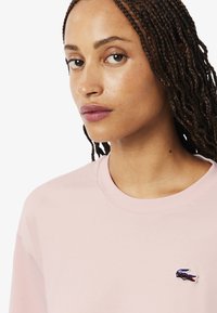 Sudadera de color rosa claro con cuello redondo, mangas cortas y un pequeño logo de cocodrilo bordado en el lado izquierdo del pecho. Textura suave.