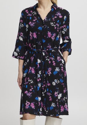 Femme portant une robe florale noire boutonnée, longueur genoux, avec des fleurs roses, bleues et blanches, cintrée à la taille par une ceinture en tissu.
