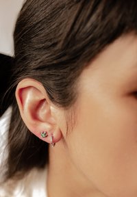 Pendiente de aro plateado con piedras preciosas multicolores y un pequeño pendiente de piedras preciosas verdes en forma de flor, exhibido en el lóbulo de la oreja.