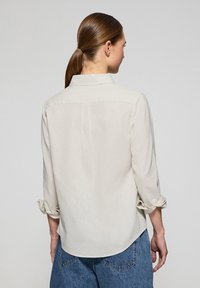 Camicia lunga beige chiaro con collo, maniche arrotolate e orlo dritto. Realizzata in un tessuto morbido e liscio con una leggera consistenza.