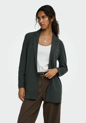 Femme aux longs cheveux foncés porte un blazer foncé, un haut blanc texturé, un pantalon marron et des colliers dorés superposés tout en regardant sur le côté.