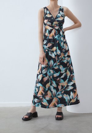 Donna che indossa un abito maxi senza maniche con scollo a V con motivo floreale multicolore e sandali neri con plateau, in piedi su un pavimento grigio.