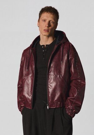 Junger Mann trägt eine burgunderrote Lederjacke mit Kapuze über einem schwarzen Hemd mit Knöpfen und schwarze Hose und steht mit den Händen in den Taschen vor einem schlichten Hintergrund.