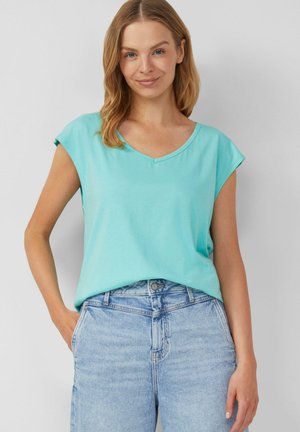 Femme aux cheveux blonds mi-longs portant un haut sans manches bleu clair à col en V et un jean taille haute bleu clair.