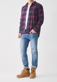 Flanellen overhemd in marineblauw en bordeauxrood ruitpatroon, wit T-shirt, lichtblauwe spijkerbroek en beige werkschoenen met gele accenten en bruine veters.