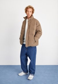 Brown puffer jacket with a high collar, zip closure, and logo detail. Paired with loose denim jeans and light sneakers with plaid accents.