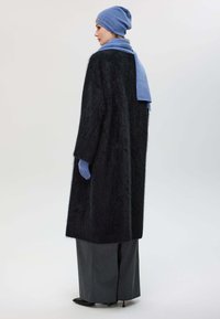 Un long manteau noir duveteux, un bonnet bleu et une écharpe, un pantalon large gris et des gants bleus. Des textures lisses et des couleurs contrastées.