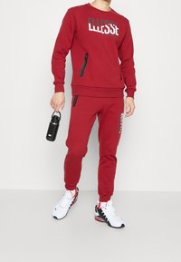 Czerwony sweter z kapturem i logo, z kieszeniami na zamek; dopasowane spodnie z nadrukiem logo. Białe buty sportowe z czarnymi akcentami oraz butelka na wodę.