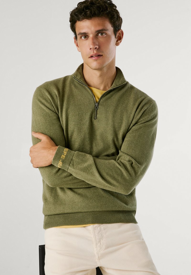 Oliwkowy sweter na zamek o fakturowanym wykończeniu, z prążkowanymi mankietami i kontrastową żółtą podszewką, noszony na żółtej koszulce.