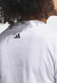 Biely bavlnený tričko s malým čiernym logom Adidas na hornej časti chrbta. Tkanina vyzerá mäkká a má hladkú textúru.