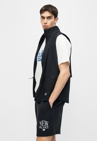 Νεαρός άνδρας που φοράει μαύρο αμάνικο γιλέκο με φερμουάρ πάνω από λευκό μπλουζάκι με γράφημα και μαύρο σορτς New Balance, με τα χέρια στις τσέπες, στραμμένος πλαγίως.