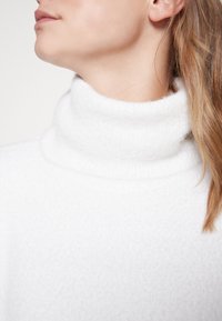 Maglione girocollo bianco, con una texture morbida e pelosa. Il colletto alto avvolge comodamente il collo, mettendo in risalto il calore del tessuto.