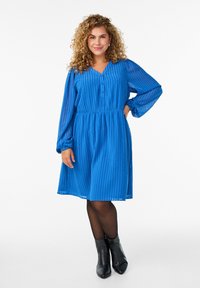 Sininen raidallinen mekko, jossa on V-pääntie, napit yksityiskohtina, pitkät hihat ja laskostettu vyötärö. Yhdistetty mustiin nilkkureihin. Tasainen, kevyt kangas.
